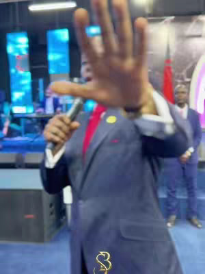 A man in a dark suit and red tie, holding a microphone and gesturing with his right hand towards the camera. His other hand holds a mic., There is crowd and lights in the background.