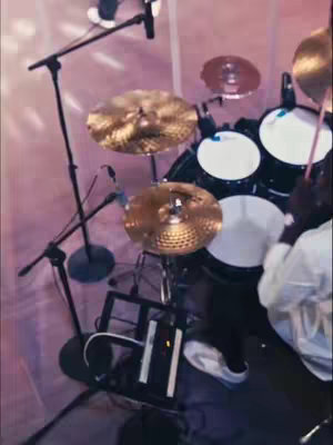 The cover image shows a drum set from an eye-level perspective. The drums are black with white drumheads. Cymbals are visible, and microphone stands are positioned to capture the audio from the drum set. A white surface is visible in the lower portion of the image, suggesting a stage or performance area. The lighting gives a soft pink hue to the scene.