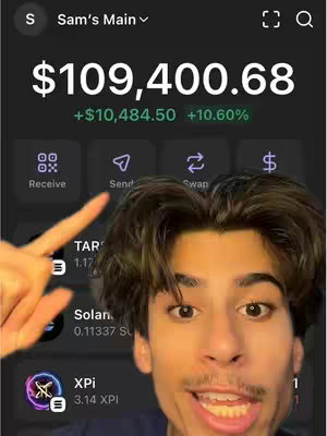 A young man is pointing at the screen of a crypto app, showing a balance of over $100,000. The background appears to be a dark app interface with various features. The image is a selfie, and the man is smiling, appearing to explain the interface.