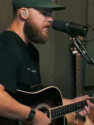 A man with a beard wearing a green cap and black t-shirt is playing an acoustic guitar and singing into a microphone. The image is well-lit and appears to be taken indoors, possibly in a studio.