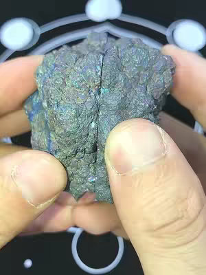 The image shows a close-up view of a crystal held between two hands against a dark background. The crystal has a rough, textured surface with a variety of colors, primarily shades of blue, green, and gray, giving it an iridescent appearance. The hands holding the crystal are fair-skinned and the focus is sharp on the crystal, with the background slightly blurred.