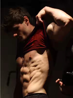 A shirtless man in a dark setting is seen pulling up his red shirt to expose his abs. The image is taken from a side angle, highlighting his toned physique. A hand is visible on the right side of the frame, interacting with the shirt. The lighting is dim, creating shadows that emphasize the muscular definition.