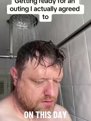 A man stands in a shower with water streaming down over his head and face. The man is looking down, with a pained expression. The background is a tiled shower, with the shower head visible nearby.