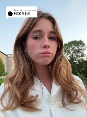 A young woman with long wavy blonde hair is centered in the frame, facing the camera. She has fair skin, subtle blush on her cheeks, and a slightly downturned expression. She's wearing a white collared shirt. A text overlay in a speech bubble appears in the top left corner. The background shows hints of trees and a building, suggesting an outdoor setting with soft lighting at dusk.