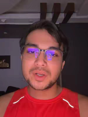 A person in a red tank top wearing glasses looks directly at the camera, shot from the chest up. The background shows an indoor setting with overhead lighting and a dark wall.