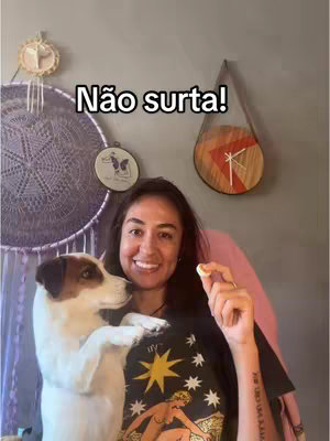 A woman with dark hair and a casual outfit smiles at the camera, holding a small object in her hand. A white dog with brown spots sits next to her, looking towards the left. In the background, there's a wall with decorative items, including a dreamcatcher and a clock.