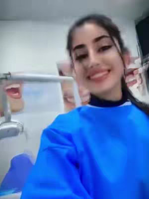 A selfie-style shot of a smiling woman in a dental setting. The woman, who has dark hair pulled back from her face and is wearing a blue top, is the focus. A dental chair and equipment are visible in the background, with a poster of teeth providing context.