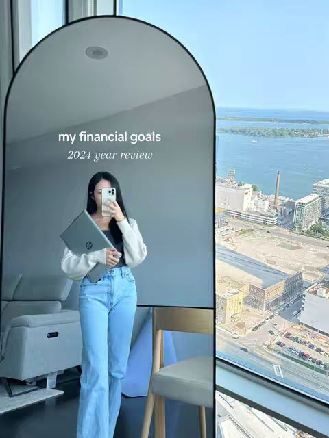 A woman is reflected in an arched mirror. The woman is holding a laptop and a phone, and wearing light wash jeans, a black top, and a white cardigan. The mirror is in front of a window that looks over a city.