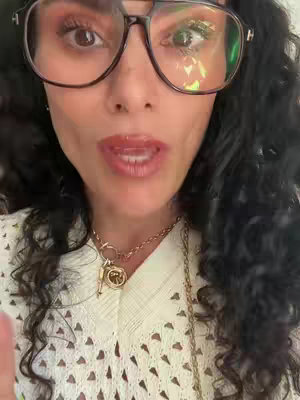 A close-up shot shows a woman with a fair complexion, wearing large-framed glasses and a white open-knit top. Her dark, wavy hair frames her face. She is looking directly at the camera, her lips slightly parted as if speaking. A gold necklace with a pendant is visible. Reflections of the light are noticeable on the lenses of her glasses. The background is softly blurred.