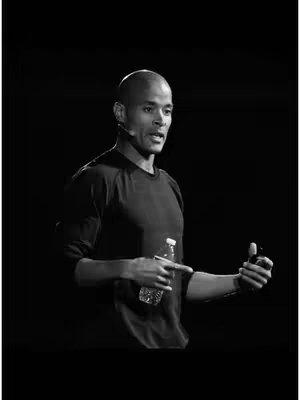 A grayscale photo shows a man giving a speech. He is bald and wears a black long-sleeve shirt, standing in front of a dark, blurred background. He holds a water bottle in his left hand and points at it with his right hand as if making a point during the speech.