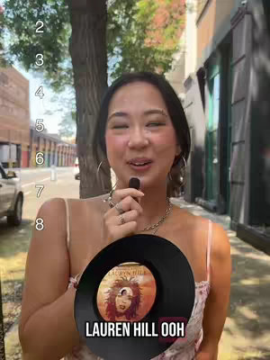 A woman stands outdoors, holding a microphone and a vinyl record. The background is a blurred street scene with a building and trees. The record is labeled 'LAUREN HILL OOH'. Numbers appear on the left side of the frame.
