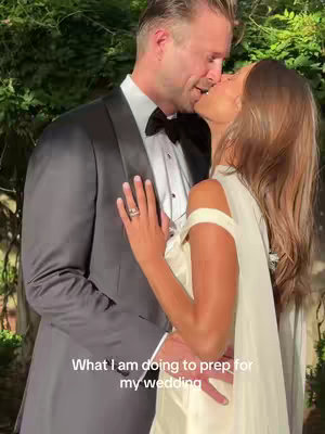 A romantic image of a couple kissing, the man in a dark tuxedo and the woman in a white wedding dress. The photo also has a blurred background of green foliage.