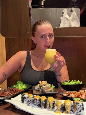 A woman sits at a table laden with sushi and other Asian-inspired dishes. She holds a tall glass of yellow liquid to drink, looking up with a slightly confused or contemplative expression. The table is filled with various plates of food, including sushi rolls with black sesame seeds, fried items, edamame, and various sauces. The background is a dimly lit restaurant setting.