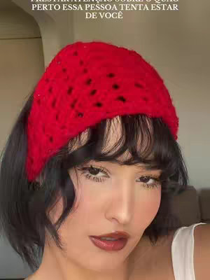 A close-up shot features a woman with fair skin. She has black hair with bangs, and is wearing a vibrant red knitted headband. Her makeup includes dark eyebrows, eyeshadow, and bold, dark red lipstick, which accentuates her facial features. The shot is well-lit. In the background, a blurred element gives a hint of an interior setting, possibly a room.