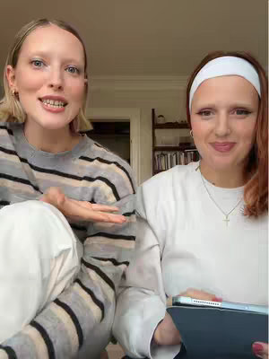 Two women are seated and facing the camera. The woman on the left is wearing a striped sweater and white pants, and she is gesturing outwards with her right hand. The woman on the right has a white headband and holds a dark tablet, smiling at the camera.