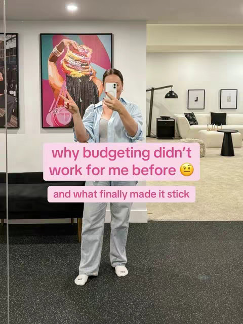 A woman is taking a selfie in a spacious room with a modern interior. She is wearing a light blue, long-sleeved shirt and light-colored pants. She is holding the phone up with the peace sign. The background includes a large colorful painting, a white couch, black side table and minimalist decor. The overlay text is in a pink box with the text: "why budgeting didn't work for me before" and then on a new line, "and what finally made it stick".