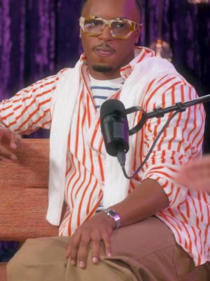 A man in a red and white striped shirt sits on a wooden bench in a studio setting. He is wearing large, square-framed eyeglasses and a white scarf. A microphone is positioned near his face. The background features a purple curtain.