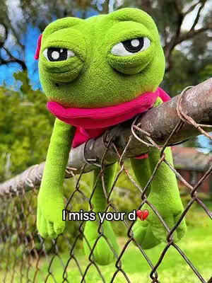 A stuffed frog with droopy eyes is perched on a metal fence. It's wearing a red scarf and has its arms and legs hanging down. Text below the frog reads, "I miss your d ❤️". The background is slightly blurred, showing trees and greenery.
