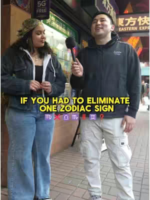 A street scene features a man and a woman standing side-by-side on a brick sidewalk. The woman is on the left, wearing a hat, denim jacket, and jeans, holding her hands in front of her. The man on the right is holding a microphone, dressed in a black hoodie and khaki pants. Behind them, a store front is visible with a partial sign that reads "FREE" and glimpses of a yellow and red sign in the background. Text overlaid at the bottom reads "IF YOU HAD TO ELIMINATE ONE ZODIAC SIGN."