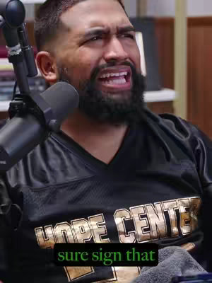 A man with a beard is the central subject in image, seen from the chest up. He is wearing a black jersey with gold lettering that reads, “HOPE CENTER.” The man has an emotional expression, indicating he might be speaking with passion or in distress, with his mouth open. A microphone is positioned close to his face. The background is blurry, showing a room with furniture and possibly bookshelves. In the bottom part of the image, there's a text overlay.