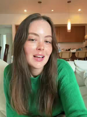 A medium shot shows a young woman with long brown hair smiling and talking to the camera. She's wearing a green sweater. The background includes a domestic interior with neutral colors.