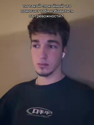 A young man with short brown hair wearing a dark sweatshirt is the focus. The image is a medium shot. Text overlayed on the image asks, "Ты такой спокойный что помогает тебя избавиться от тревожности?" (You're so calm that it helps you get rid of anxiety?). The background is softly lit, suggesting an indoor setting.