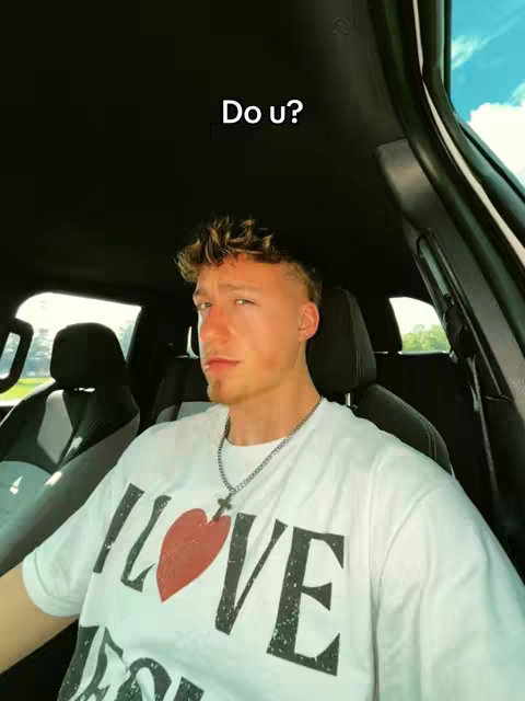 A young man, with light skin and short, styled blonde hair, is positioned inside a car. He's wearing a white t-shirt with the words "I LOVE" above a red heart and the word "VE" below it. The background shows the inside of the car with the car seat, dashboard and windshield visible. The text "Do u?" is overlaid on the top of the image where the image shows a cropped version of the creator.