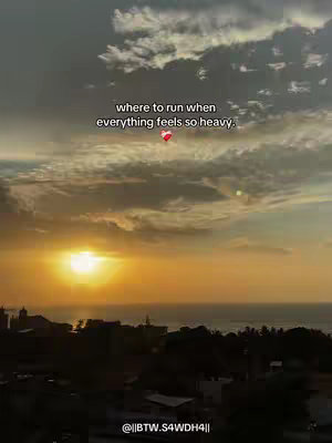 A scenic image of a sunset over the ocean. The sky is filled with clouds painted in shades of grey, orange, and yellow. In the foreground, there's a silhouette of buildings and trees. Text overlayed on the image reads, 'where to run when everything feels so heavy.' with a heart emoji.