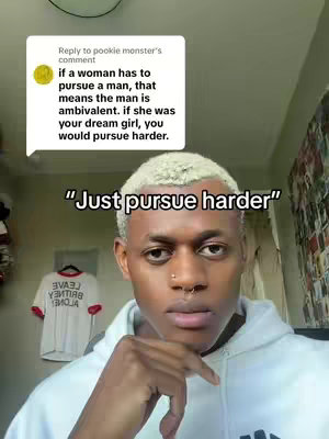 A young man with light hair and a nose ring is centered in a medium shot. He wears a white hoodie. To the left, a speech bubble contains text, and behind him, a shirt hangs on a rack. The background is a cluttered room.