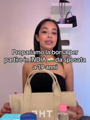 A woman in a black top holds a cream-colored tote bag in front of a blurred background. On the bag, the word "HT" is visible. The overlay text reads: Preparariamo la borsa per partire in INDIA ๐ฎ๐ณ da sposata a 19 anni. Blue and white and pink items are visible in front of the woman.