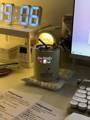 A well-lit, indoor shot of a desk setup, featuring a modern digital clock at the top with blue LED numbers indicating the time, a white lamp, and a light green Snoopy-themed tumbler with a black straw placed on a white cushion. The desk is covered with papers with handwritten notes. The color palette is warm and inviting, with soft yellow lighting.