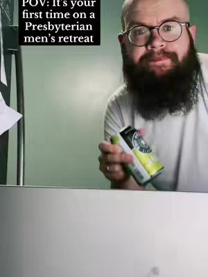 A man with glasses and a beard faces the camera, holding a beverage. Text overlayed on the top left says 'POV: It's your first time on a Presbyterian men's retreat'. The background is a simple, light green wall. The man is wearing a white t-shirt. The composition is a medium shot, focusing on the man and a portion of the surrounding environment.