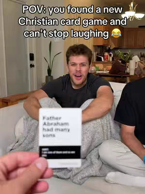 A man with short brown hair sits on a white couch, holding a white card. The card reads 'Father Abraham had many sons' with text below it. The man is smiling and looking up and to the camera. The background shows a casual living space. The text overlay reads 'POV: you found a new Christian card game and can't stop laughing'.