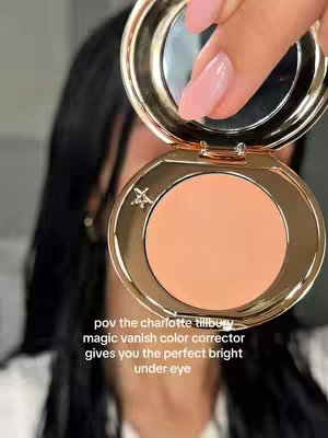 The image shows a close-up of a woman's face, holding a gold compact with a peach-colored concealer. The woman has long dark hair and well-manicured nails. The text overlay reads "pov the charlotte tilbury magic vanish color corrector gives you the perfect bright under eye."