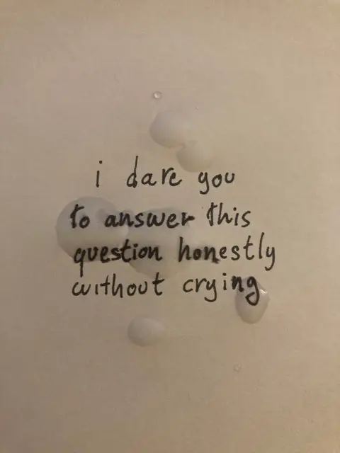 A minimalist image of a white surface, possibly paper or a wall, with the handwritten text 'i dare you to answer this question honestly without crying'. There are several translucent, irregular-shaped droplets of a clear liquid scattered around the text.