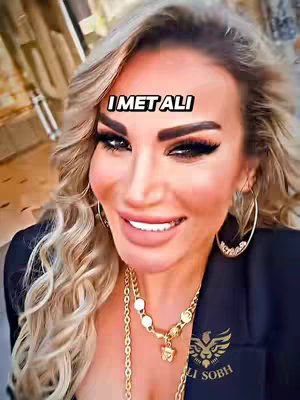A selfie-style shot of a woman with blonde, wavy hair, wearing heavy makeup, and gold jewelry. The background is blurred, suggesting an indoor setting with a hint of architectural details.