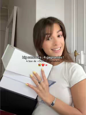 A young woman with long brown hair holds up a stack of white gift boxes in front of her chest. She smiles at the camera and has yellow nail polish. Text overlays state a contest to give away boxes of products.