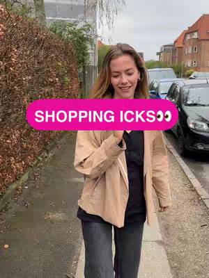 A young woman walks toward the camera on a paved path with a street and buildings in the background. She's wearing a tan jacket over a black top and dark pants. A pink banner with white text covers a portion of her face.