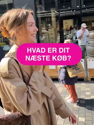A woman smiles and looks to the left. She wears a light-colored trench coat and has her hair pulled back with a decorative clip. A pink oval with white text floats near her face. Behind her, a window and a street scene provide a blurred background, with other people walking.