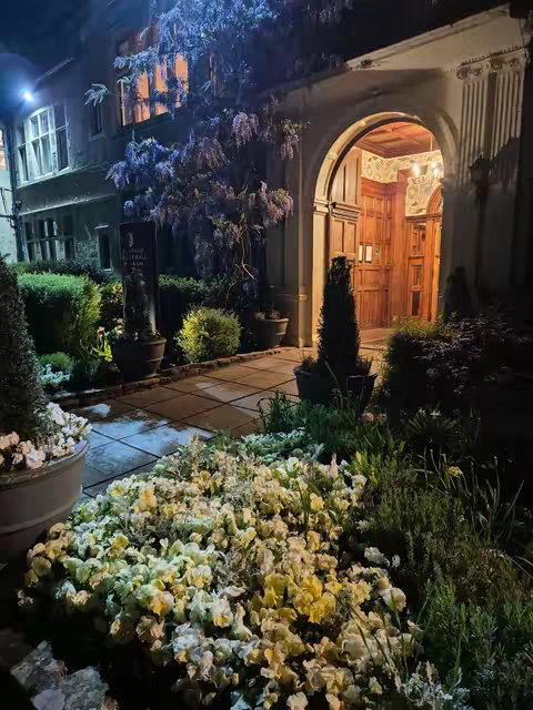 An exterior shot of an upscale building entrance at night. The entrance is framed by an arched doorway with wooden doors, and illuminated by warm, inviting light from within. The foreground is filled with a variety of plants and flowers.