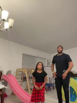 A casual indoor shot featuring a man and a young girl. The man, positioned on the right, is wearing a black t-shirt and black pants, and has his hands on his hips. The girl, on the left, is wearing a black top and a red plaid skirt. In the background, there's a pink slide next to a wooden climbing frame