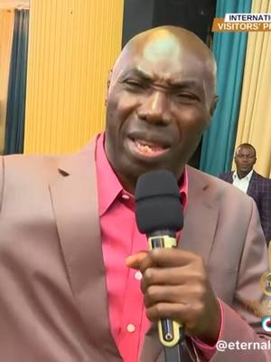 A medium shot shows a man with a shaved head, wearing a salmon-colored suit jacket over a bright red shirt. He holds a microphone close to his mouth, with a slightly pained or intense expression. In the background, there's a blurred crowd and a banner that says 'INTERNATIONAL VISITORS PLATFORM'.