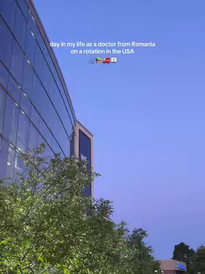 A daytime shot of a modern building with a glass exterior. Green trees are in the foreground, with a clear blue sky as a backdrop. Text overlayed in white describes the video content as a doctor's life in the USA. Multiple emojis are shown on top too.