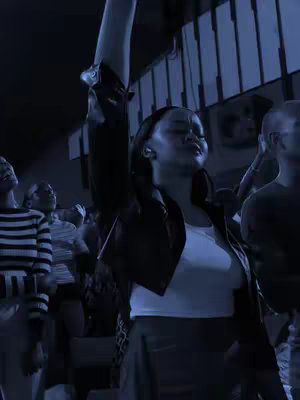 A dimly lit image with a blue tint shows a group of people in what appears to be a worship setting. A woman in the foreground, wearing a white top and black jacket, has her arm raised. Other individuals in the background are looking up. The lighting has a cool tone.