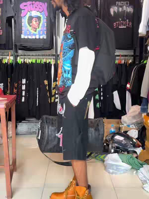 A man stands in a clothing store wearing a graphic tee with short black shorts, boots, and a backpack. The background features racks of clothes.