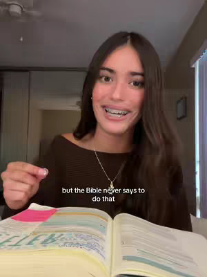 A young woman with long brown hair and braces is smiling and looking directly at the camera while holding a Bible opened in front of her. The top of a room with a ceiling fan and a window are in the background.