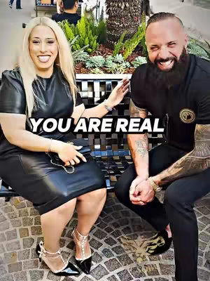 The cover image presents two people, a woman and a man, sitting on a park bench. The woman is on the left, with long blonde hair, wearing a black dress. The man is on the right, with a full beard and wearing a casual black outfit. The scene is brightly lit, suggesting an outdoor setting.