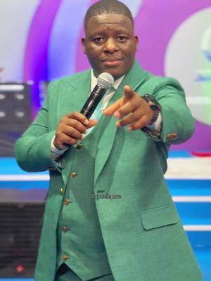 A man in a green suit holds a microphone and points his right index finger directly at the camera, indicating a message, in what seems like a studio setting with light background. He is of African descent, smiling, and appears to be in the midst of a speech or presentation.