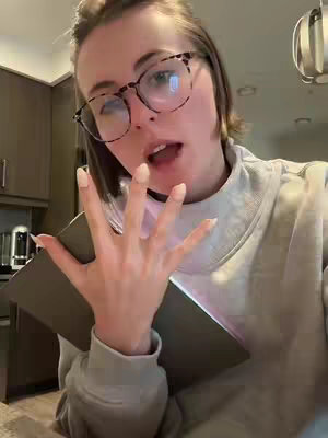 A woman with light skin, wearing glasses and a light-colored sweater, holds a tablet-like device with her hand open in front of her face. The overhead lighting reflects off the glasses. The background shows a kitchen space with cabinets.