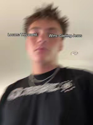 A blurred, low-angle shot features a young man with short, messy brown hair, looking slightly upwards. He's wearing a black t-shirt with an emblem on it and a silver chain. Overlaid text reads "Losses: This world." to the left and "Wins: Gaining Jesus" to the right.